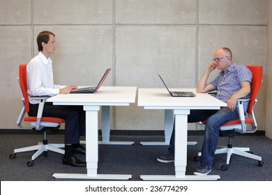 Man Working In Correct Sitting Posture In Office Watching His Colleague Sitting In Bad Position At Workstation