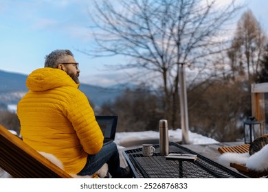 Man working from cozy cabin in mountains, sitting on terrace with laptop, enjoying cup of coffee. Concept of remote work from peaceful location. Hygge at work. - Powered by Shutterstock