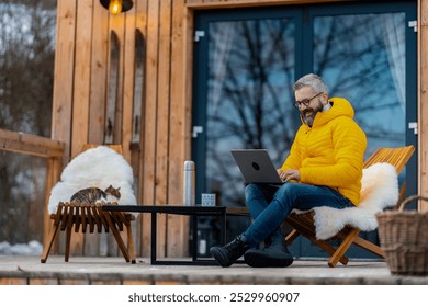 Man working from cozy cabin in mountains, sitting on terrace with laptop, enjoying cup of coffee. Concept of remote work from peaceful location. Hygge at work. - Powered by Shutterstock