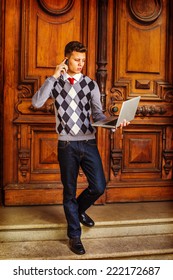 Man Working Hard. Wearing A Patterned Sweater, Red Tie, Blue Jeans, Leather Shoes, A Young Guy Is Waling Down From Office, Talking On His Mobile Phone, Working On A Laptop Computer In The Same Time.