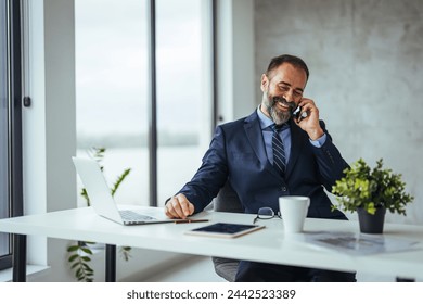 Man working at his desk at office. Smiling mature businessman holding smartphone sitting in office. Middle aged manager ceo using cell phone mobile apps and laptop. Digital technology applications  - Powered by Shutterstock