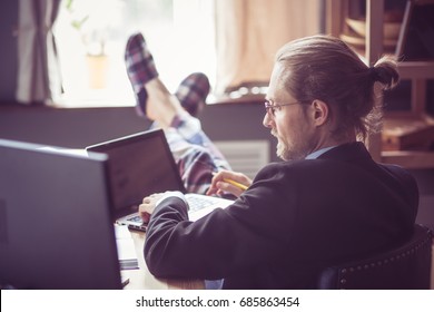 Man Working At Home Office. Freelancer Working With Computer And Laptop With His Feet In Slippers On Wooden Table. Toned Image.