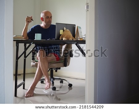 Man working from home sitting at table wearing underpants holding a pen and using computer seen through doorway in blurred foreground    