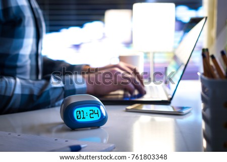 Man working late. Workaholic or being behind schedule concept. Business person in modern office building or home at night using laptop. Time in digital clock on table in workstation. 