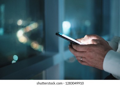 Man Working In Modern Office Late At Night. Busy Caucasian Manager At Work Texting On Cell Phone, With City And Traffic Lights In Background. Close-up Of Hands Holding Smartphone