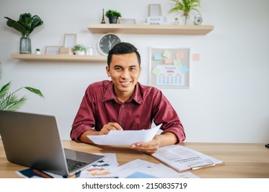 man working in office holding papers with laptop on desk