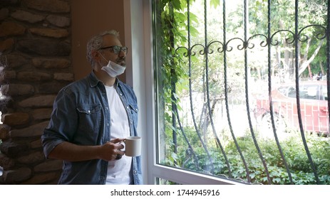 Ein im Büro arbeitender Mann sieht mit Maske und Kaffee aus dem Fenster.
