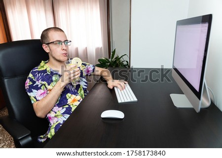 Man working on the computer while enjoying drinking a refreshing summer drink, wearing a hawaiian shirt. Successful young man, influencer working from home