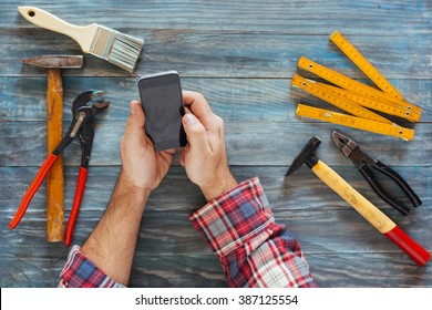 Man Working On A DIY Project With His Phone, Wood Shavings And Carpentry Tools And Construction All Around, Top View