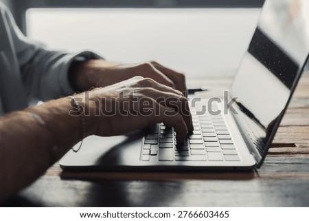 Man working on a laptop at a rustic wooden desk, typing with focused hands in natural light; conveys remote work, productivity, technology and professional concentration in a casual office setting.