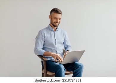 A man working on a laptop while sitting on a wooden chair in a bright, minimalistic office during daytime hours