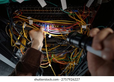 Man working on network server, holding flashlight and connecting cables inside server rack, focusing on maintenance and troubleshooting in data center environment