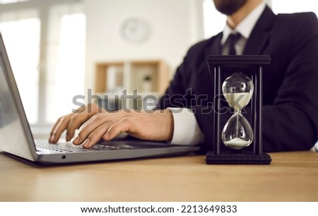 Man working on notebook computer at desk with hour glass. Employee typing on PC keyboard and trying to meet deadline. Don't waste time, it's a limited resource. Carpe diem. Seize the day. Seize moment
