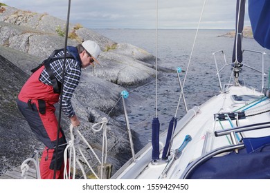 Man Working On Untie Rope Sailboat Out From Harbour Shore Island Rock In Sweden Over Summer Sky Background.