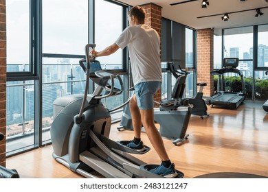 Man working out on an elliptical trainer in modern gym with city view. Male doing cardio training on elliptical machine in fitness studio in residental building. Fitness, wellness, healthy lifestyle - Powered by Shutterstock