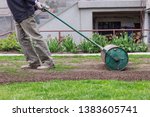 Man in working outfit pulls lawn roller behind. Necessity after long winter and spring for flat surface. Landscaping on the garden. Summer worker