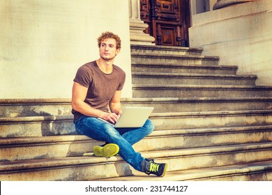 Man Working Outside. Wearing Dark Brown T Shirt, Blue Jeans, Black Sneakers, A Young Sexy Guy With Curly Hair Is Sitting On Stairs Outside Office, Crossing Legs, Thinking, Typing On Laptop Computer. 