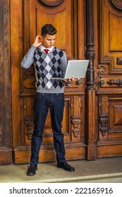 Man Working Outside. Wearing A Patterned Sweater, Red Tie, Blue Jeans, Leather Shoes, A Young Guy Is Standing Outside Office, Looking Down, Scratching Head, Thinking, Working On A Laptop Computer.