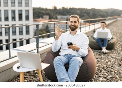 Man working remotely on rooftop using phone and laptop while showing OK gesture. Woman in background using laptop. Outdoor workspace for remote work, digital nomad lifestyle. - Powered by Shutterstock