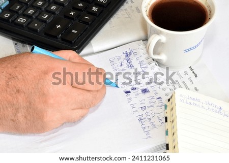 A man is working at a table. He writes something in a notepad. Nearby is a calculator and a cup of coffee.