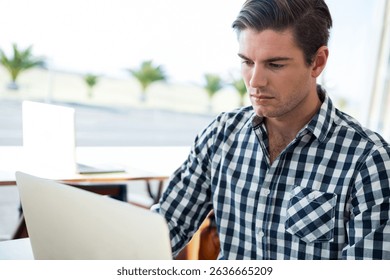 Man working at table in café on silver laptop near windows with potted palms, copy space. Co-working, freelance, modern, minimalistic, urban, professional, creative