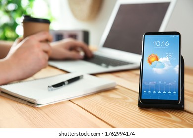 Man working at table and smartphone with open weather forecast app indoors