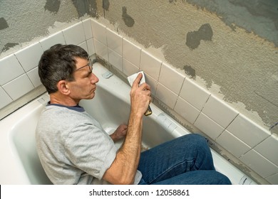 Man working in a tight space while applying ceramic tile to a bathtub enclosure wall. Viewed looking down.