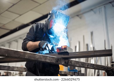 A man working as a welder in a protective mask and work clothes performs work with a MIG-MAG welding machine. Production of welding works in a factory.