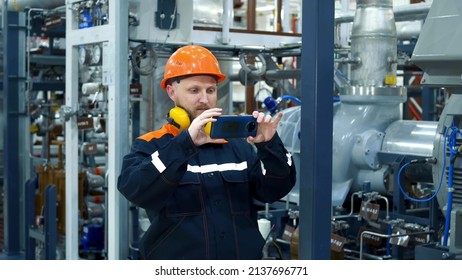 The Man In The Workplace. A Bearded Man In A Helmet Controls Equipment Using A Smartphone In A Gas Compression Shop. Noisy Work Space. Oil And Gas Industry