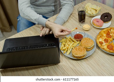 A man works at a computer and eats fast food. unhealthy food: Burger, sauce, potatoes, donuts,chips. Unhealthy concept.