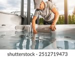 A man works diligently at a hot tub, checking the water quality while surrounded by greenery in a bright, sunny backyard.