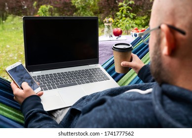 Man Works On Laptop In The Garden Sitting On Hammock And Using Phone And Drink Coffee. Empty Screen Of Laptop And Mobile With Mock Up. Home Office With Relax And Nature. Freelancer Outdoors. Close Up.