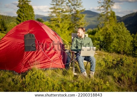 Man works on laptop while traveling with tent in the mountains. Charging computer with portable solar panel hanging on tent. Concept of remote work at campsite and renewable energy
