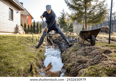 Man in workwear clearing a waterlogged garden path in early spring with a shovel and wheelbarrow amidst a rural landscape.