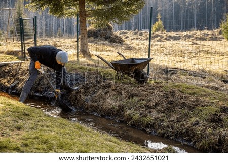 Man in workwear clearing a waterlogged garden path in early spring with a shovel and wheelbarrow amidst a rural landscape.