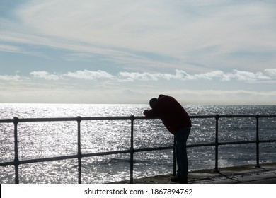 A man wrapped up against the cold leans on iron railings on the shore. He faces out to sea and is hunched over with his head on his arms looking sad. Shot against the light for silhouette. 