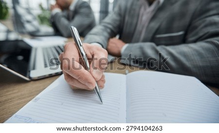 Man writes notes in notebook at desk. Co-worker rests hand on head, focused. Office environment feels professional and calm. Ideal for business, education, or meeting themes