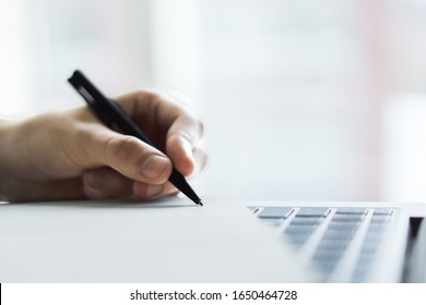Man writes with a pen in diary on laptop keyboard in a sunny office, business and education concept. Close up