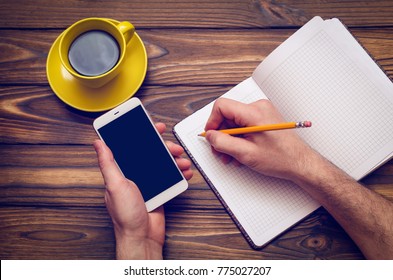 A man writes something in a notebook with a yellow cup of coffee and a phone on a wooden table.