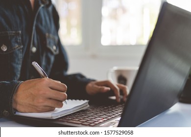 Man Is Writing A Document In Office Room With A Black Laptop And Window. A Businessman Wearing Jeans Is Analyzing Business Concept Data.