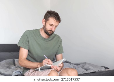 Man writing in notebook while sitting on unmade bed, focused on morning plans and thoughts, starting day with quiet journaling routine in simple home interior