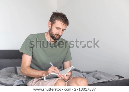 Man writing in notebook while sitting on unmade bed, focused on morning plans and thoughts, starting day with quiet journaling routine in simple home interior
