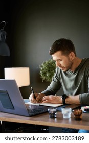 Man writing notes in notebook at home desk near laptop and smartphone in warm interior light. Concept of planning, remote work and productivity
