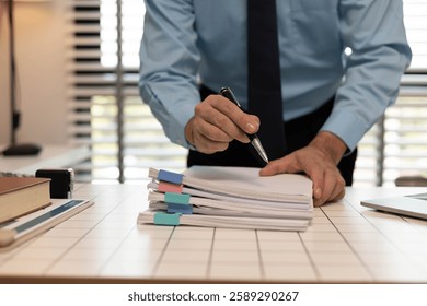 A man is writing on a stack of papers with a pen. The papers are organized in a neat stack on a table. The man is focused on his work, possibly preparing a report or a document
