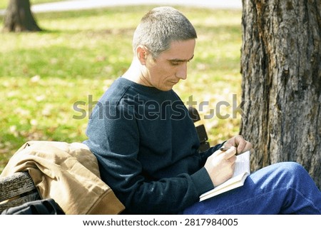 Man writing in small notebook while relaxing beneath tree in public park during calm autumn day. Represents offline reflection, mindful journaling, cognitive reset, and sustainable attention management habits.