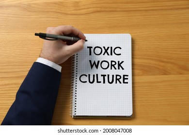 Man Writing Text Toxic Work Culture In Notebook At Wooden Table, Top View