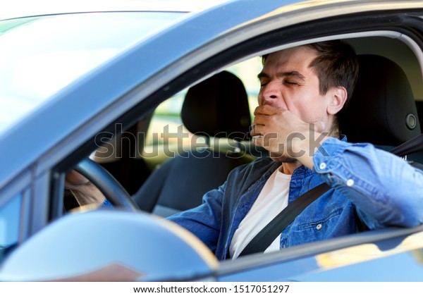 Man is\
yawning behind steering wheel of car. Sleepy driver is riding in\
automobile. Male is violating rules of road. Driving without rest\
concept. Deadly danger on route of modern\
city.