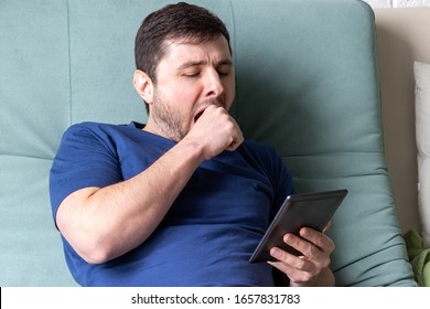 Man Yawns While Reading An E-book. A Resting Male In A Blue T-shirt Sits On A Green Sofa With An Electronic Book In One Hand, Yawns, Covering His Mouth With His Second Hand