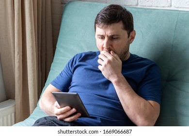 Man Yawns While Reading An E-book. A Resting Male In A Blue T-shirt Sits On A Green Sofa With An Electronic Book In One Hand, Yawns, Covering His Mouth With His Second Hand