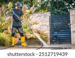 A man in yellow boots operates a pressure washer, cleaning a patio area surrounded by greenery and a garden gate on a bright afternoon.